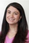 Head-and-shoulders portrait of a smiling woman with long dark hair, wearing a pink top and small earrings, against a plain light background.