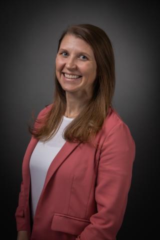Mairin Batten smiling in front of a gray background wearing a white shirt and a pink blazer.