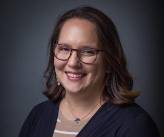 A woman with brown hair and glasses smiles against a blue background