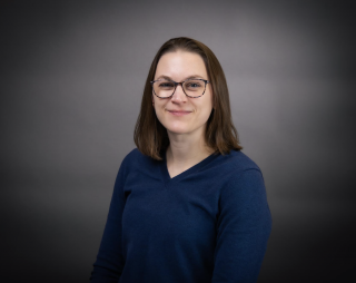 Portrait of Clare Gallagher smiling slightly at the camera against a dark gray studio background, wearing dark-rimmed glasses and a navy blue V-neck sweater, with straight shoulder-length brown hair.